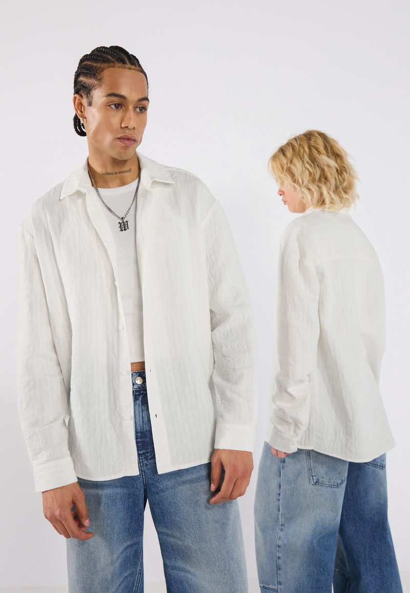 Chemise blanche texturée à ouverture frontale avec un col, des manches longues et des détails de boutons visibles, associée à un jean large bleu clair.
