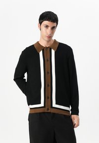 Svart kavaj med brun krage, prydd med vita och bruna vertikala ränder. Texturerad tyg och fem svarta knappar framtill.