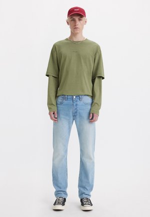 Camiseta verde de gran tamaño con un pequeño logo, jeans rectos de color azul claro y zapatillas negras. Una gorra roja completa el atuendo informal.