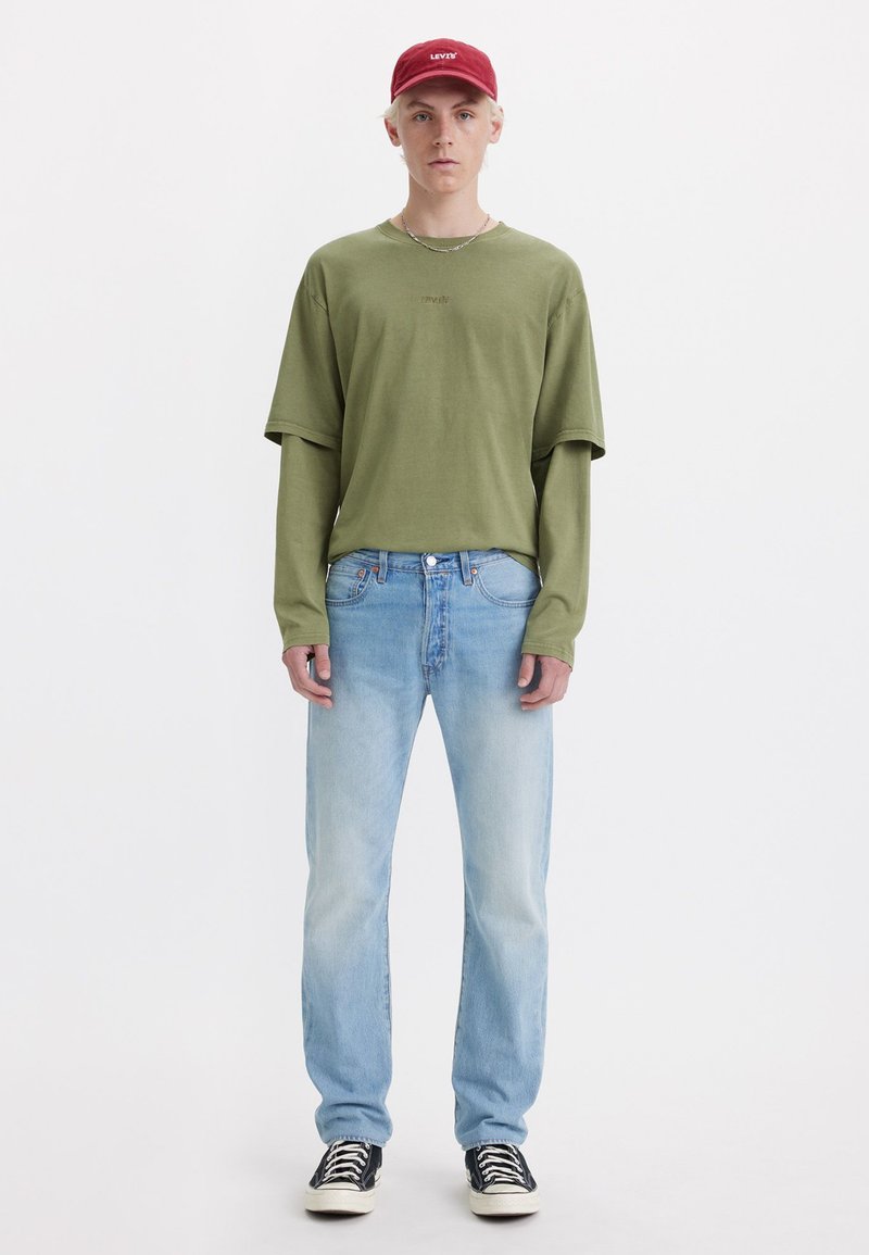 Zelena oversize majica s malim logom, svijetloplave ravne traperice i crne tenisice. Crvena kapa upotpunjuje ležerni outfit.