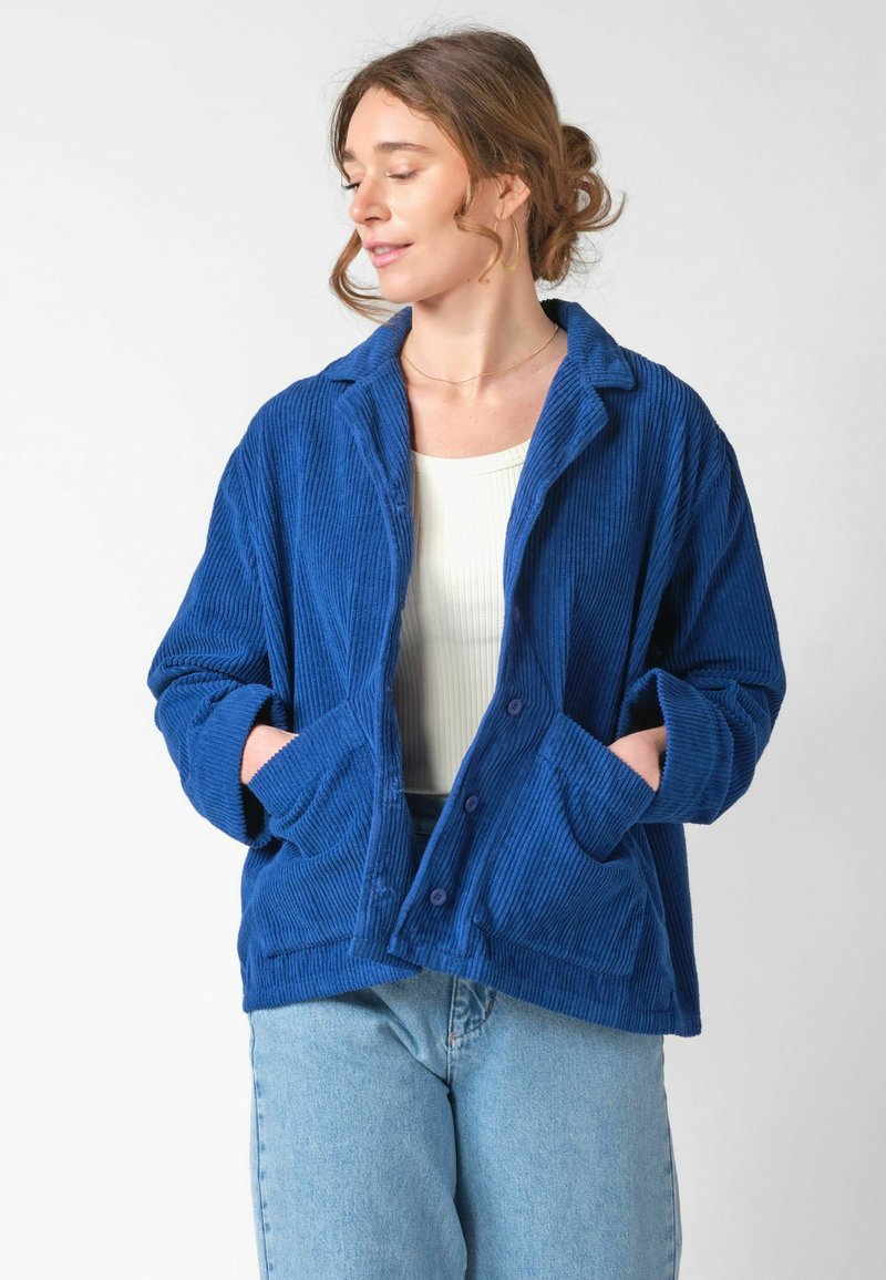 Femeie cu părul prins într-un coc lejer, purtând o jachetă albastră din catifea peste un top alb și blugi deschis la culoare, cu mâinile în buzunarele jachetei, uitându-se spre stânga.