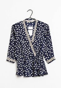 Blouse marine avec motif de cœurs blancs, encolure en V, bordure en dentelle le long des bords, manches longues, et détail de taille nouée. Suspendue à un crochet noir.