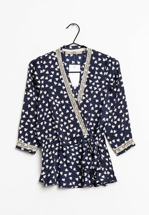 Blouse marine avec motif de cœurs blancs, encolure en V, bordure en dentelle le long des bords, manches longues, et détail de taille nouée. Suspendue à un crochet noir.