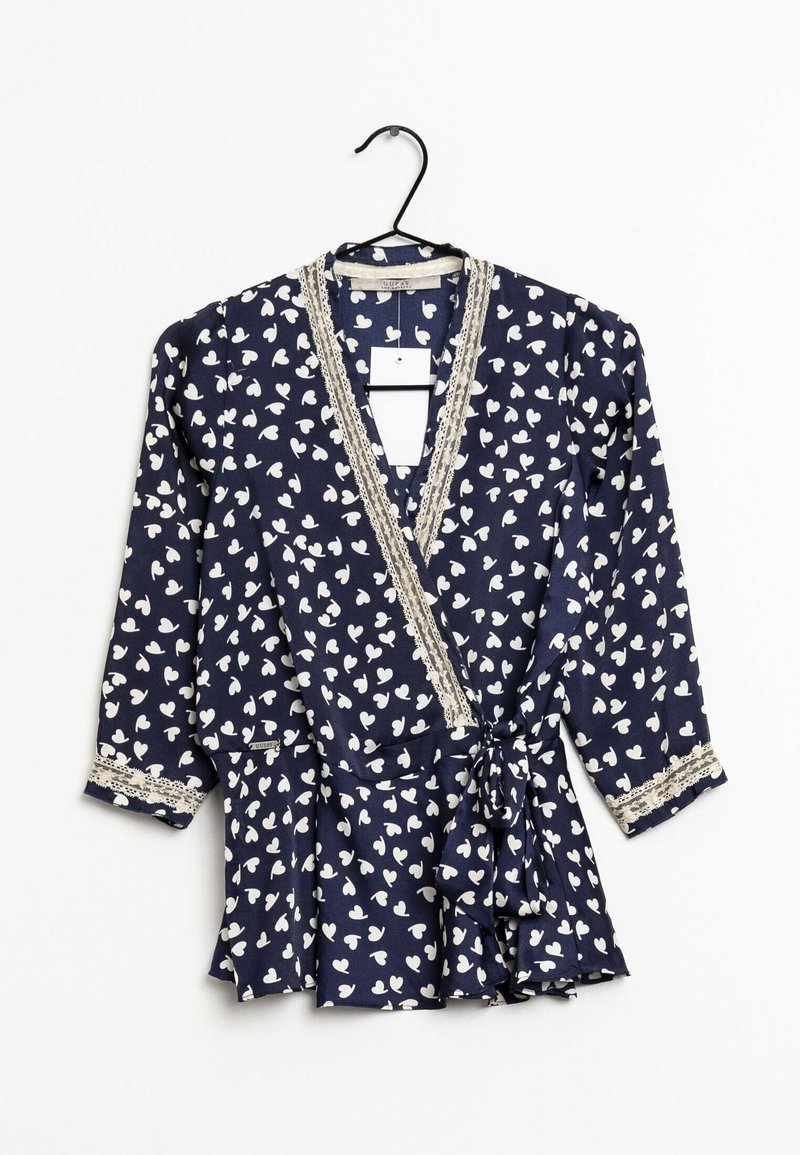 Blouse marine avec motif de cœurs blancs, encolure en V, bordure en dentelle le long des bords, manches longues, et détail de taille nouée. Suspendue à un crochet noir.