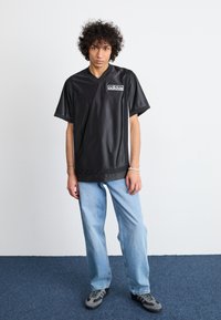Camiseta atlética preta, de manga curta e decote em V, com um logotipo no lado esquerdo do peito. Combinada com jeans de perna reta azul clara e ténis cinzentos.