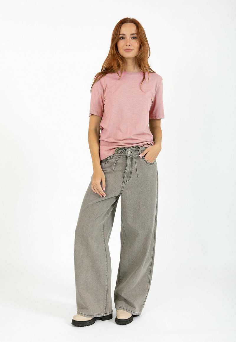 Jeune femme aux longs cheveux roux, portant un t-shirt rose, un jean gris à jambes larges et des bottes beige, debout avec une main dans la poche.