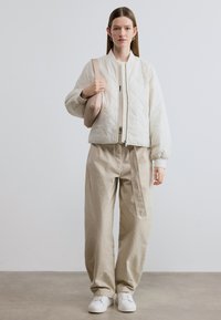 Λευκό μπουφάν μπόμπερ με καπιτονέ ύφασμα, μπεζ παντελόνια με φαρδιά πόδια και λευκά αθλητικά παπούτσια. Συνοδεύεται από μια απαλό ροζ τσάντα χιαστί.