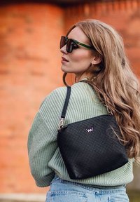 Černá texturovaná kabelka s geometrickým vzorem, s krátkým popruhem a lesklým logem, nošená s zeleným svetrem a modrými džíny.