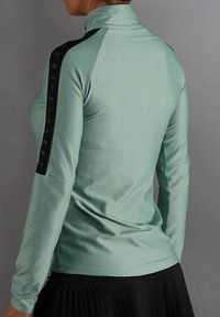 Mintgroen shirt met lange mouwen, een gestructureerde afwerking, hoge kraag en zwarte tape-details op de linkermouw met witte tekst.