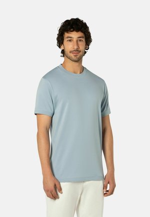 Man met kort krullend haar, gekleed in een lichtblauw shirt met korte mouwen en witte broek, staand tegen een effen lichte achtergrond.