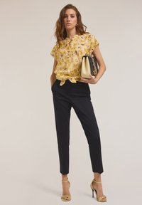 Blouse florale jaune à manches courtes avec détail noué, associée à un pantalon noir ajusté et des sandales à talons jaunes. Sac à main noir et or.