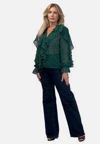 Green chiffon blouse with ruffled details, long sleeves, paired with dark flared jeans. Model stands against a neutral background.