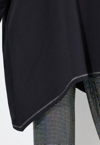 Zwarte tuniek met een gebogen zoom en stikdetails, gecombineerd met glanzende, meerkleurige leggings met een stippenpatroon.