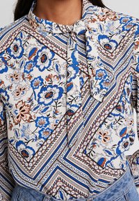 Květinová halenka s krémovým pozadím, zdobená modrými a oranžovými květy, geometrickými vzory a límcem se zavázanou mašlí.