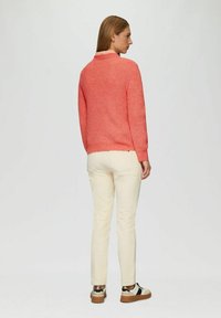 Korallenfarbener Strickpullover mit rundem Ausschnitt, kombiniert mit off-white Slim-Fit-Hosen und Sneakers. Die Textur ist leicht grob und verleiht Tiefe.