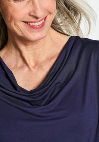 Cowl-Neck-Top aus dunkelblauem Stoff mit sanften Drapierungen und glatter Textur, die Hauttöne unterhalb des Ausschnitts hervorhebt.