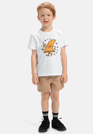 T-shirt blanche avec un graphique de taco cartoon orange, texte "Vagues Savoureuses". Shorts beiges, chaussures noires avec des chaussettes rayées blanches et vertes.