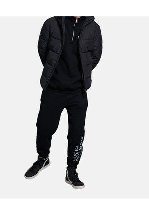 Duvet noir matelassé avec col zippé, sweat-shirt noir et pantalon de survêtement noir avec texte blanc sur la jambe gauche. Baskets noires.