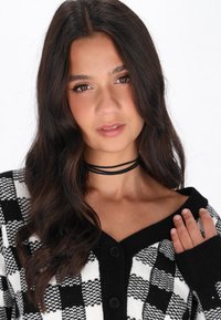Cardigan a scacchi bianco e nero con scollo a V, maglia strutturata, quattro bottoni e un choker nero indossato attorno al collo.