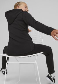 Felpa nera con cappuccio abbinata a pantaloni, seduta su una sedia bianca in metallo. Presenta maniche lunghe, una texture liscia e una vestibilità comoda.