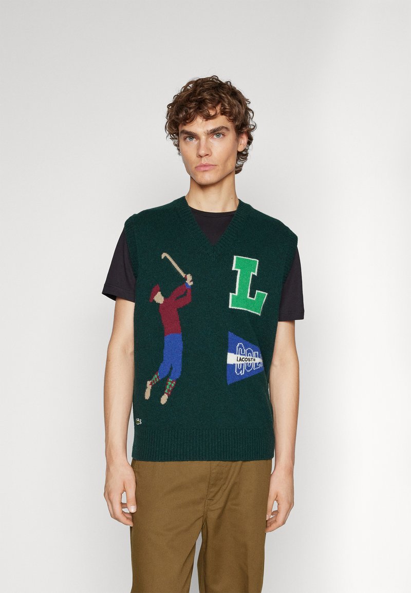 Giovane uomo che indossa un gilet a maglia verde scuro con una grafica di un golfista, una grande "L" verde e il logo Lacoste sopra una maglietta nera e pantaloni marroni.