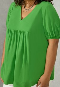 Blusa verde a maniche corte con scollatura a V, dettagli arricciati e tessuto morbido e leggero con una superficie liscia.