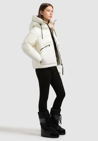 Hvid pufferjakke med hætte, med sorte lynlåse og detaljer. Sammen med sorte leggings og knæhøje sorte støvler. Glat tekstur.