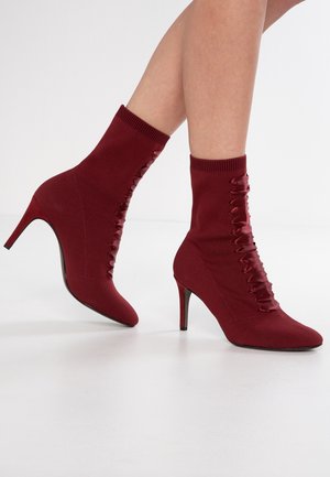 Bordowe botki do kostki z ostrymi noskami, szpilkami i ozdobnym sznurowaniem po bokach, noszone na gołych nogach na białym tle.