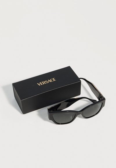 Μαύρα ορθογώνια γυαλιά ηλίου με σκούρα φακούς δίπλα σε μαύρη θήκη Versace με χρυσό λογότυπο σε λευκή επιφάνεια.