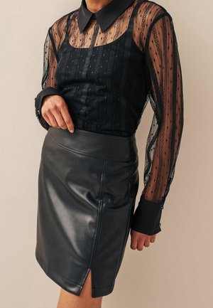 Femme portant un chemisier en dentelle noire transparente avec col et poignets, associé à une jupe en cuir noire avec une fente devant.