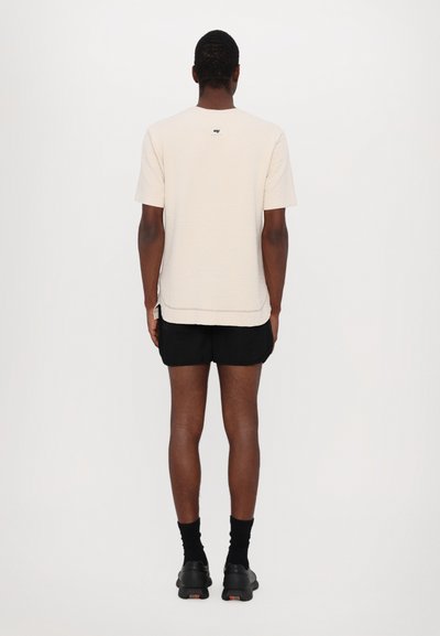 Lichtbeige textuur korte mouwen shirt met zijsplitten, gedragen met zwarte shorts en zwarte schoenen, gezien vanaf de achterkant.