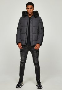 Dunkelgraue Puffjacke mit Kapuze aus Kunstfell, gestepptem Muster und Logo-Details. Tragen mit zerrissenen schwarzen Jeans und Turnschuhen.