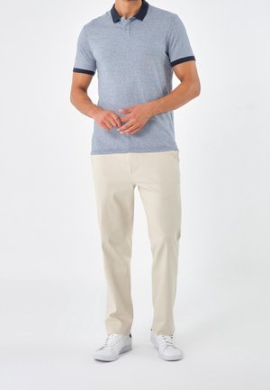 Mand iført en lyseblå polo med mørk krave og beige bukser, der står med hænderne foldet, iført hvide sneakers.