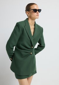 Vihreä bleiseri, jossa on muotoiltu malli, kultaiset nappiyksityiskohdat ja sileä pinta, yhdistettynä beigeen ribattuine kaulukselliseen paita ja shortseihin.