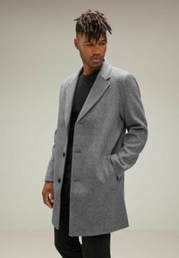 Abrigo de lana gris con solapas de muesca, frente de botones y bolsillos laterales; usado sobre una camisa negra, con tejido texturizado y un corte entallado.