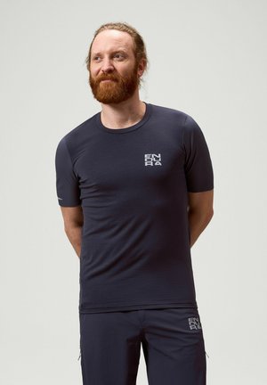 Man met rood haar en baard, die een marineblauw Endura T-shirt en korte broek draagt, staat met zijn handen achter zijn rug en kijkt naar links.