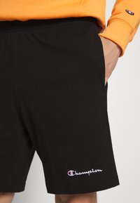 Pantalones cortos deportivos negros, fabricados con una tela suave, con una cintura elástica, bolsillos laterales y un logo bordado en blanco de "Champion".