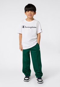 Bílá bavlněná tričko s logem "Champion" v tmavě modré barvě, doplněné zelenými tepláky s bílými bočními pruhy a logem, a tmavými teniskami.