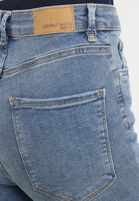 Takakatsomo sinihapuisista farkuista, joissa on yksi takatasku ja vaalea etiketti, jossa lukee "perfect jeans ginatricot."