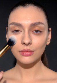 A makeup brush with a gold handle is held against the cheek of a person with light skin, wearing natural makeup and has dark hair.