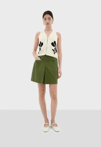 Crème gebreide vest met zwart bloemenpatroon, gecombineerd met een groene plissérok. Model draagt witte instapschoenen en staat tegen een neutrale achtergrond.