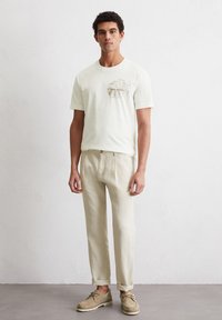 Cream t-shirt with embroidered design on the chest, paired with light linen trousers and beige shoes. Simple, casual design with a relaxed fit.