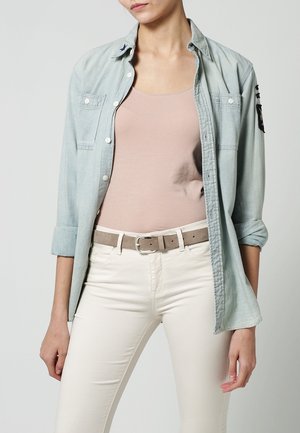 Femme portant une chemise bleue claire ouverte sur un débardeur beige, avec un pantalon blanc et une ceinture beige, debout devant un fond uni.