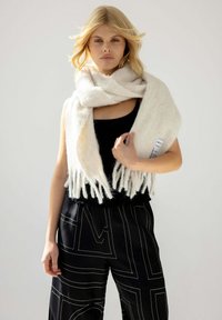 Écharpe blanche et duveteuse avec des franges, enroulée autour d'une personne, associée à un haut noir et un pantalon noir texturé avec des lignes géométriques blanches.