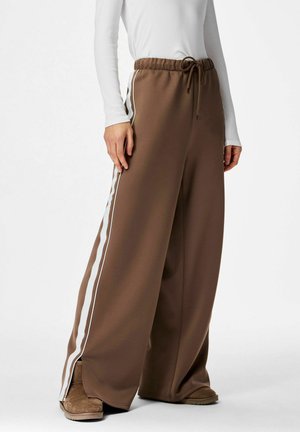 Personne portant un pantalon marron à jambes larges avec des bandes blanches sur les côtés, un haut blanc à manches longues et des bottes marron, debout devant un fond neutre.