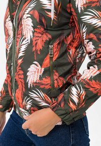 Vihreä pommitusmallinen takki, jossa on punavalkoinen trooppisten lehtien kuosi, vetoketjukiinnitys, joustavat hihansuut ja kiristysnauhallinen huppu.