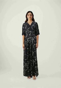 Ženska v črni maksi obleki z belim abstraktnim risanjem obraza, stoji ob enobarvni svetli ozadju, opremljena z uhani in sandali.