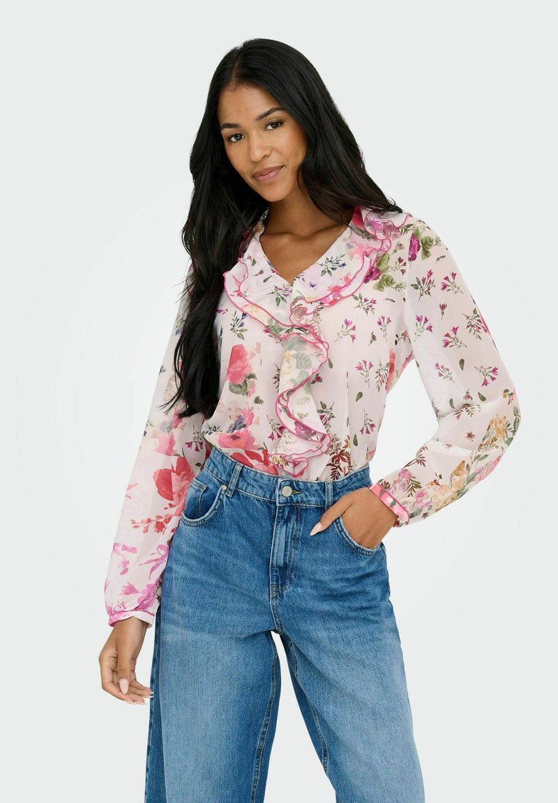 Blusa floral en rosa claro con escote y puños con volantes, que presenta patrones de flores multicolores. Llevada con jeans de tiro alto en azul.