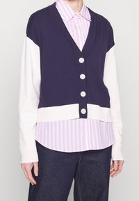 Mörkblå och krämfärgad blockfärgad cardigan med djup V-ringning, prydd med fem stora knappar, bärs över en rosa randig skjorta.