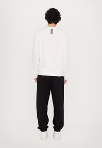 Hvid sweatshirt med rund halsudskæring, synlige sømme og sorte striber på ryggen. Sammen med sorte løstsiddende bukser og hvide sneakers.